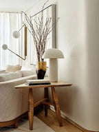 A Clérachic table lamp on a three-legged wooden side table beside a vase of branches and an arm lamp with a white globe.