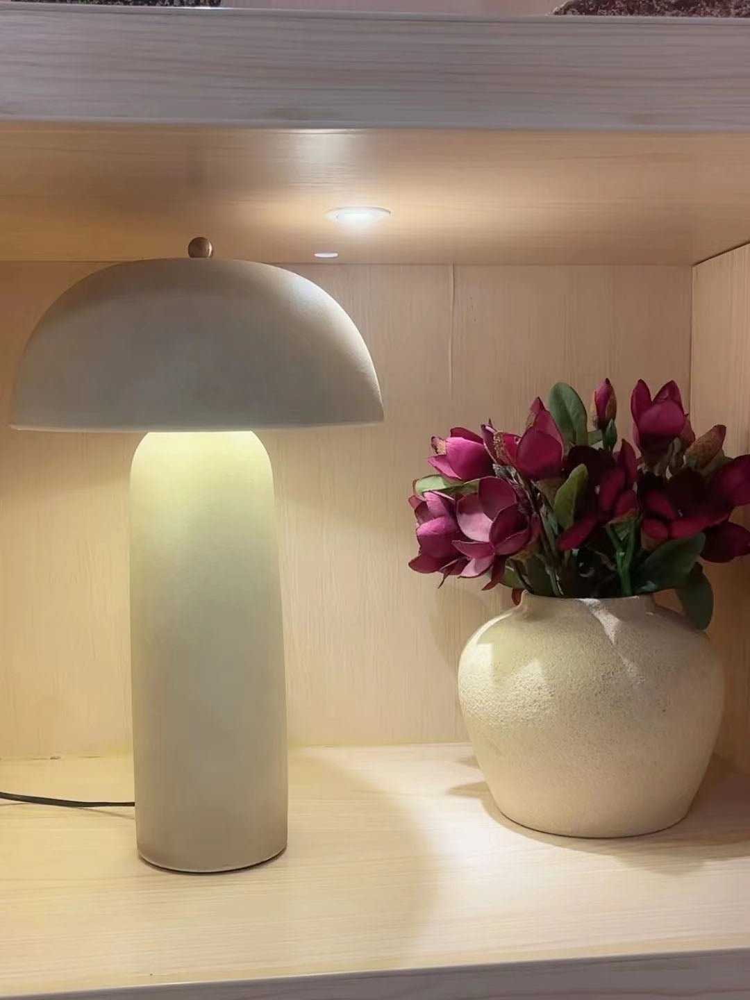 Lit lamp with a glowing, pale green-grey column on a shelf beside a cream vase of magenta flowers; a Clérachic design.