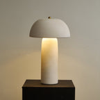 Glowing column and domed shade on a dark plinth, with a warm pool of light on the wall behind; a Clérachic table lamp.