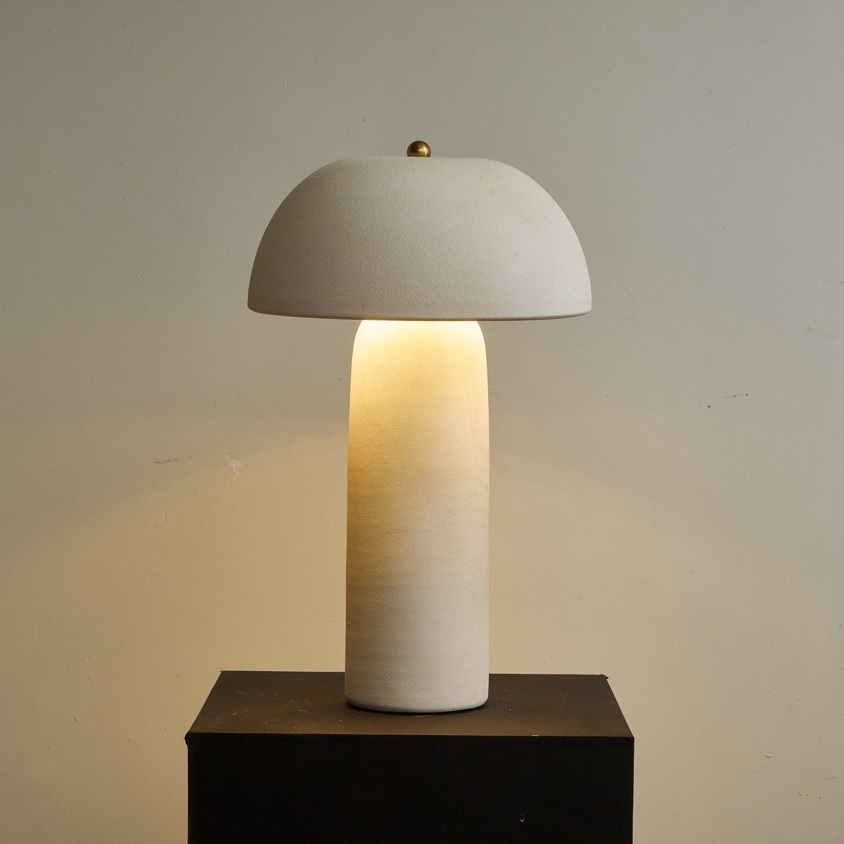 Glowing column and domed shade on a dark plinth, with a warm pool of light on the wall behind; a Clérachic table lamp.
