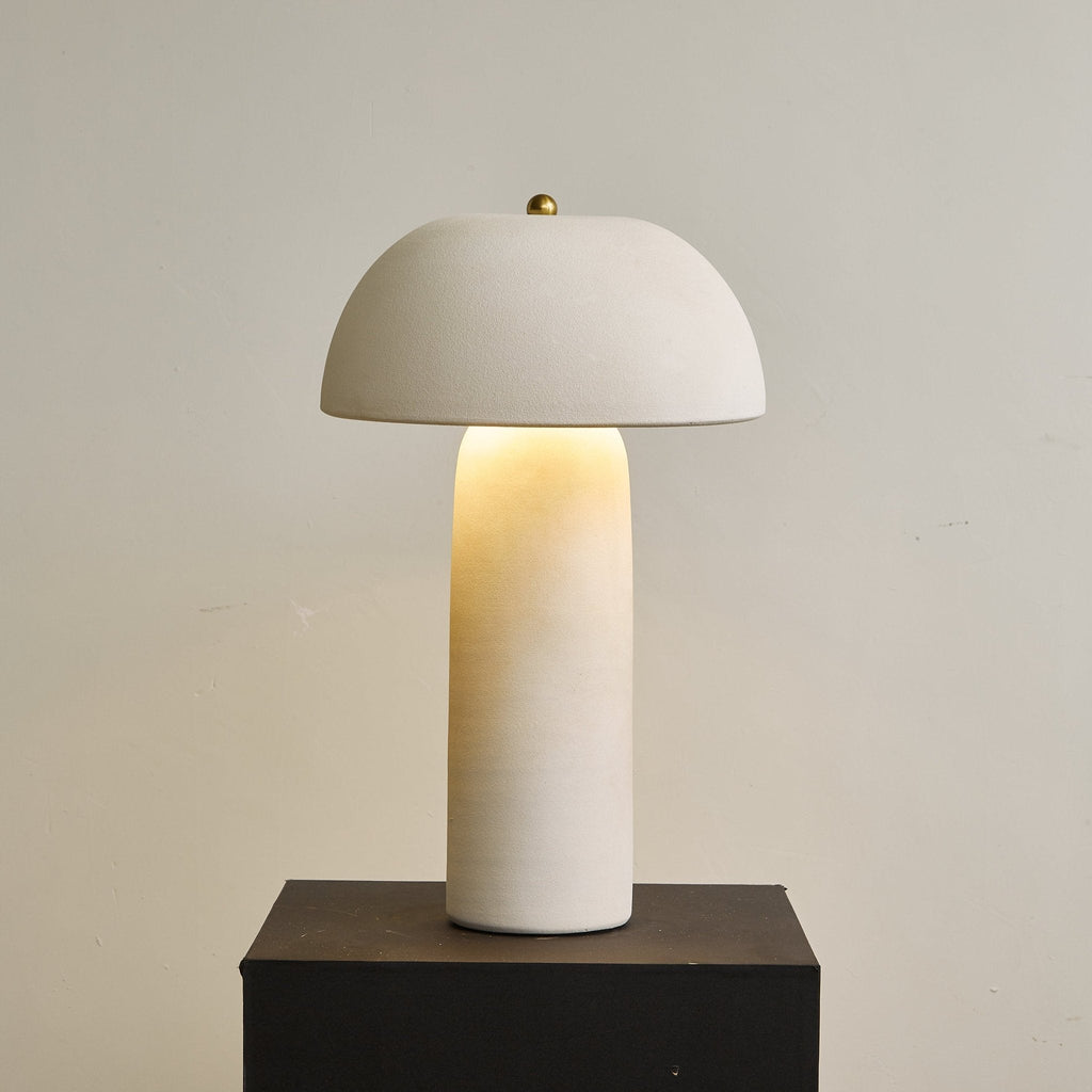 Straight-on view of the lit lamp on a black plinth, with a brass finial on the dome and a beige wall behind; from Clérachic.
