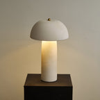 Centred view of the lit lamp on a black plinth, in front of a plain greige wall; this Clérachic table lamp.