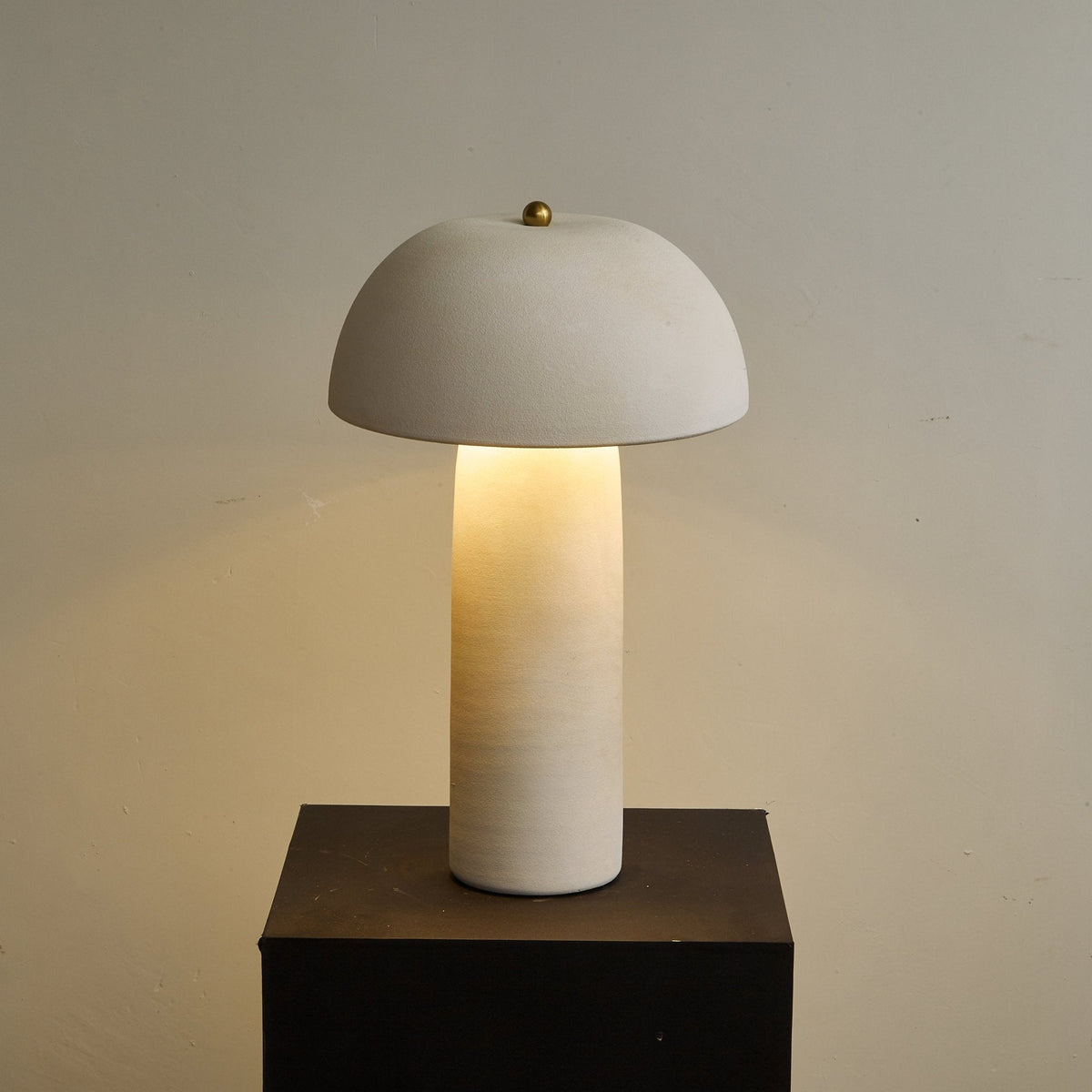 Centred view of the lit lamp on a black plinth, in front of a plain greige wall; this Clérachic table lamp.