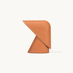 A Clérachic table lamp in orange on a plain white backdrop, with a cone base and a folded shade tilting forward over it.