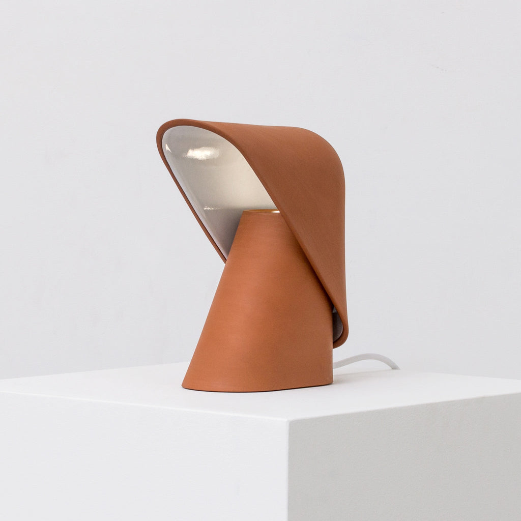 Orange Clérachic table lamp on a white plinth, its folded shade open to show a pale inner surface and a white cord running behind.