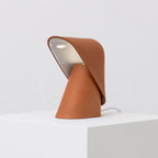 Orange Clérachic table lamp on a white plinth, its folded shade open to show a pale inner surface and a white cord running behind.