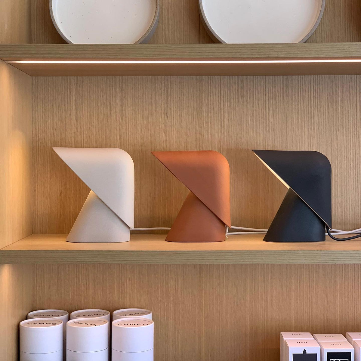 White, orange and black versions of this Clérachic table lamp side by side on a light wood shelf, above stacked white tubes.