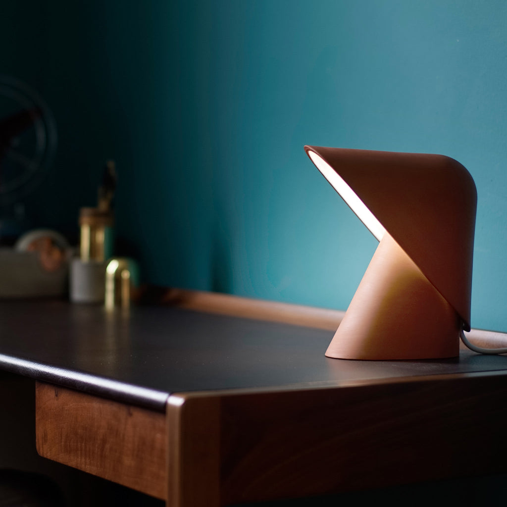 This Clérachic table lamp in orange casts light from under its folded shade on a dark wooden desk, with a teal wall behind.