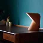 This Clérachic table lamp in orange casts light from under its folded shade on a dark wooden desk, with a teal wall behind.