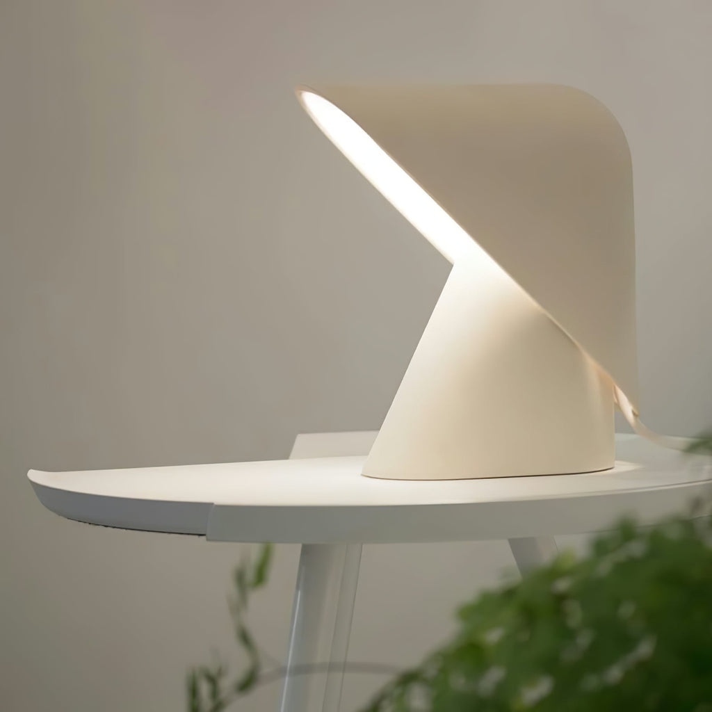 Lit white Clérachic table lamp on a small round white table, its folded shade throwing a strip of warm light, with a plant at the edge.