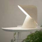 Lit white Clérachic table lamp on a small round white table, its folded shade throwing a strip of warm light, with a plant at the edge.