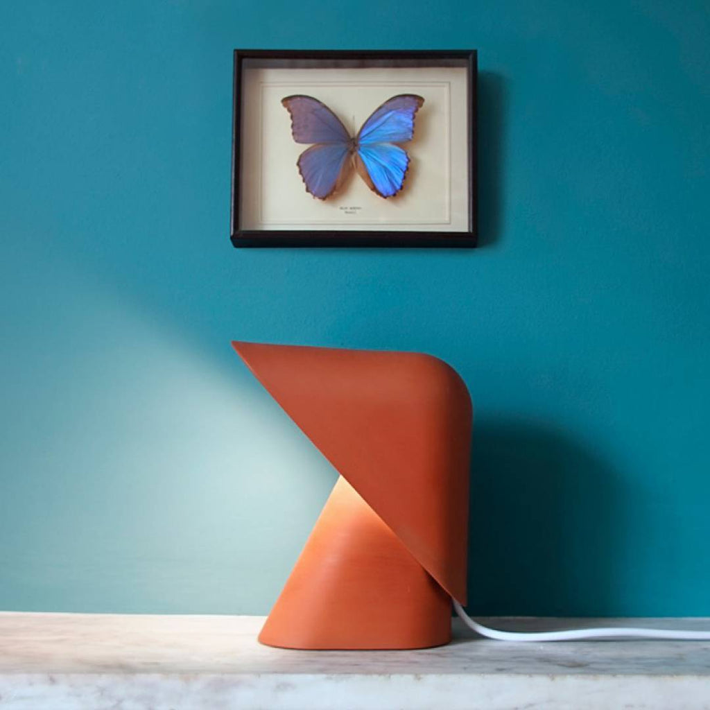 Orange Clérachic table lamp on a marble shelf against a teal wall, with a framed blue butterfly hung above it.