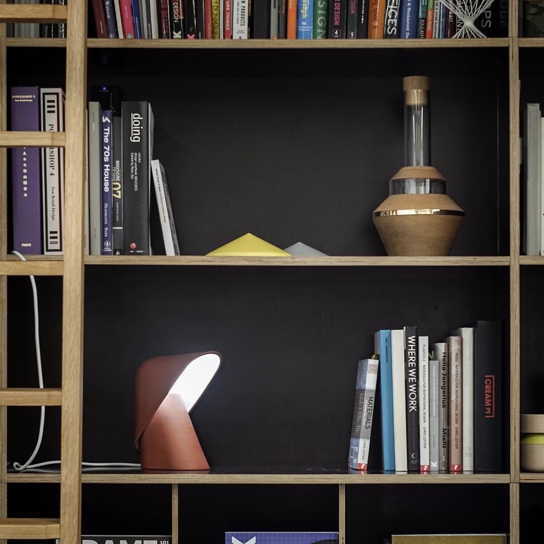 Small orange Clérachic table lamp lit on a lower bookshelf shelf, among books and a glass bottle on a dark backdrop.