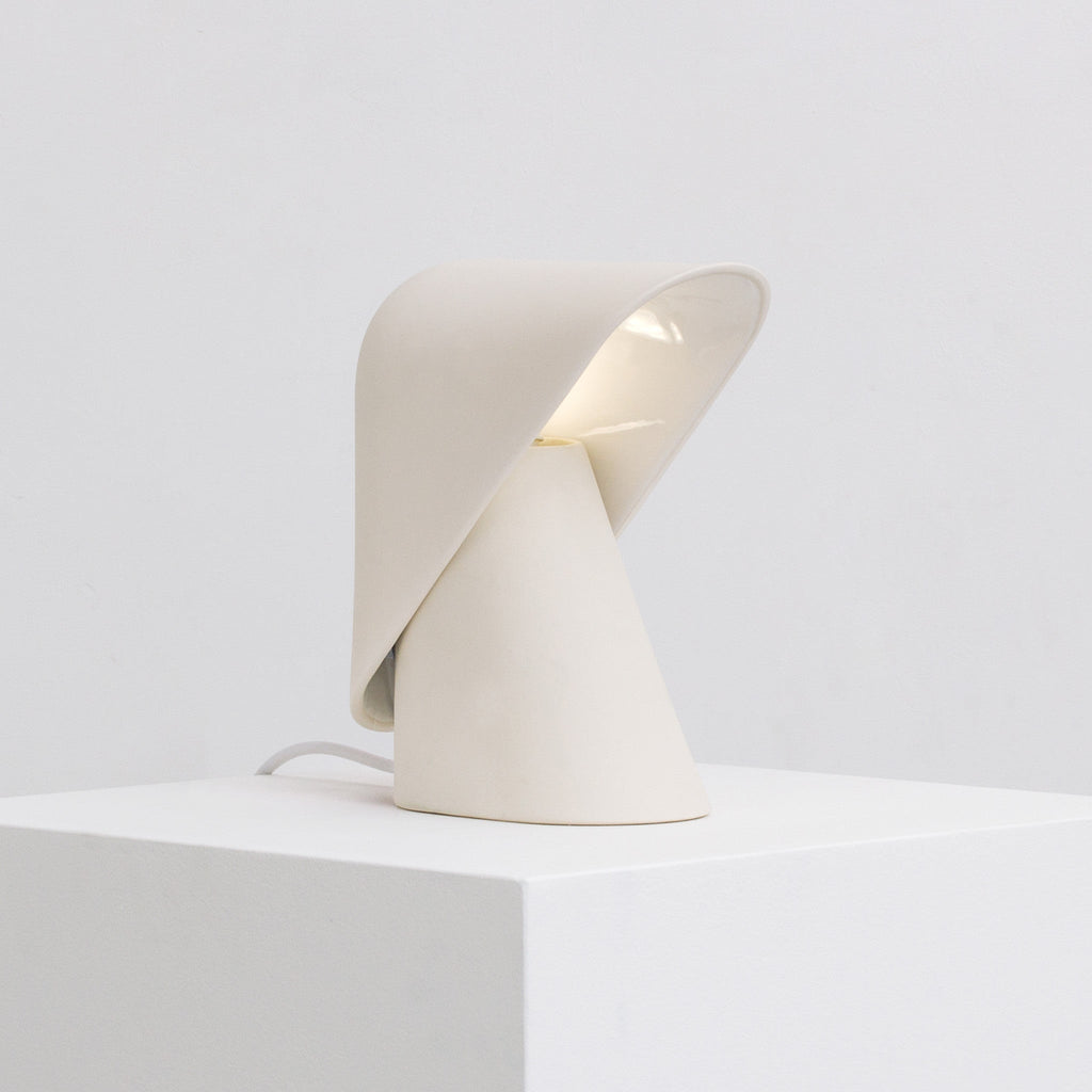 White Clérachic table lamp on a white block, with the folded shade lifted to show its pale lit interior and a cord at the left.