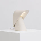 White Clérachic table lamp on a white block, with the folded shade lifted to show its pale lit interior and a cord at the left.