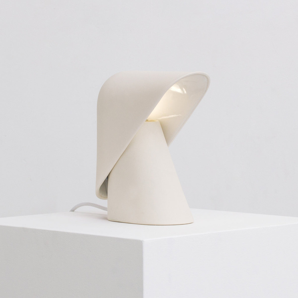 White Clérachic table lamp on a white block, with the folded shade lifted to show its pale lit interior and a cord at the left.