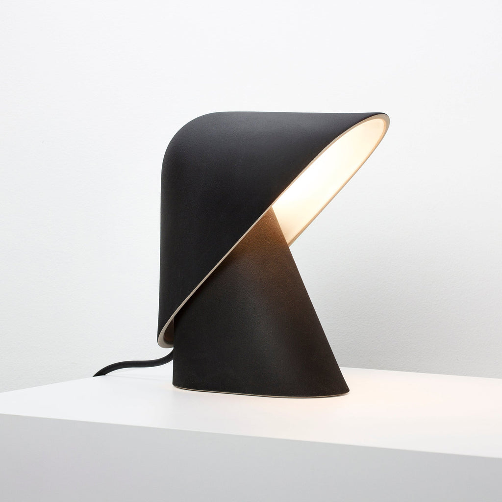 Black Clérachic table lamp on a white surface, with a cream-lined fold in the shade and a soft glow inside it.
