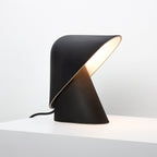 Black Clérachic table lamp on a white surface, with a cream-lined fold in the shade and a soft glow inside it.