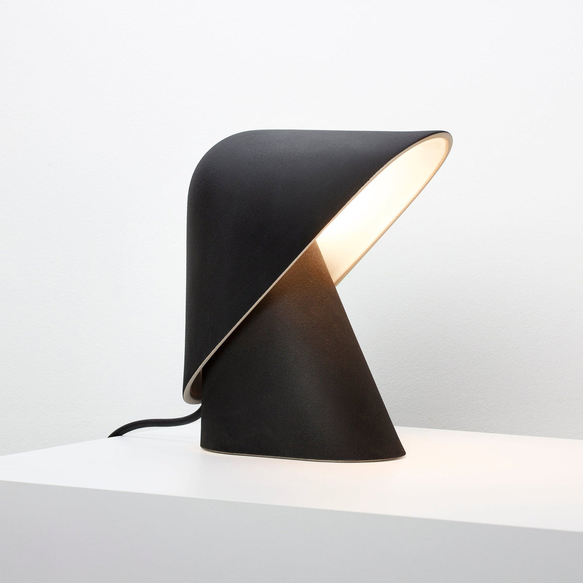 Black Clérachic table lamp on a white surface, with a cream-lined fold in the shade and a soft glow inside it.