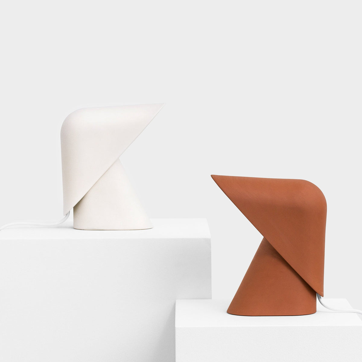 A white and an orange Clérachic table lamp on stepped white blocks against a pale grey wall.