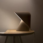 Dark Clérachic table lamp lit on a round wooden side table, its shade fold glowing and a cord trailing to the right.