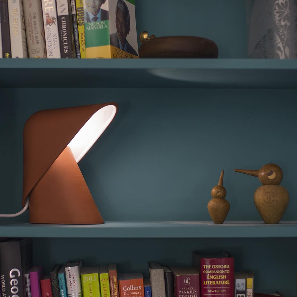 Orange Clérachic table lamp lit on a teal shelf, with two small wooden bird figures at the right and rows of books above and below.