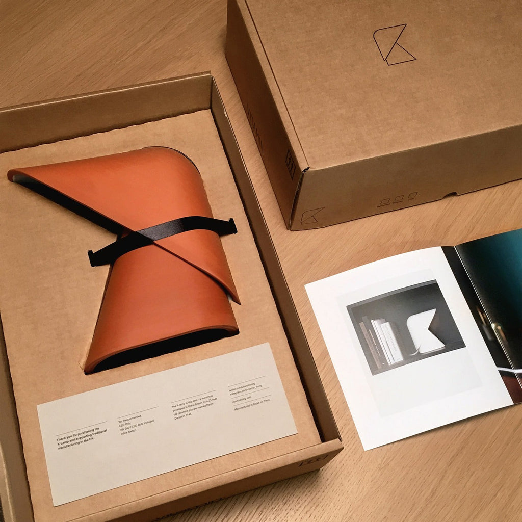 Open cardboard box holding an orange Clérachic table lamp in a soft cover with a black strap, beside a printed card and a booklet.