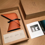Open cardboard box holding an orange Clérachic table lamp in a soft cover with a black strap, beside a printed card and a booklet.