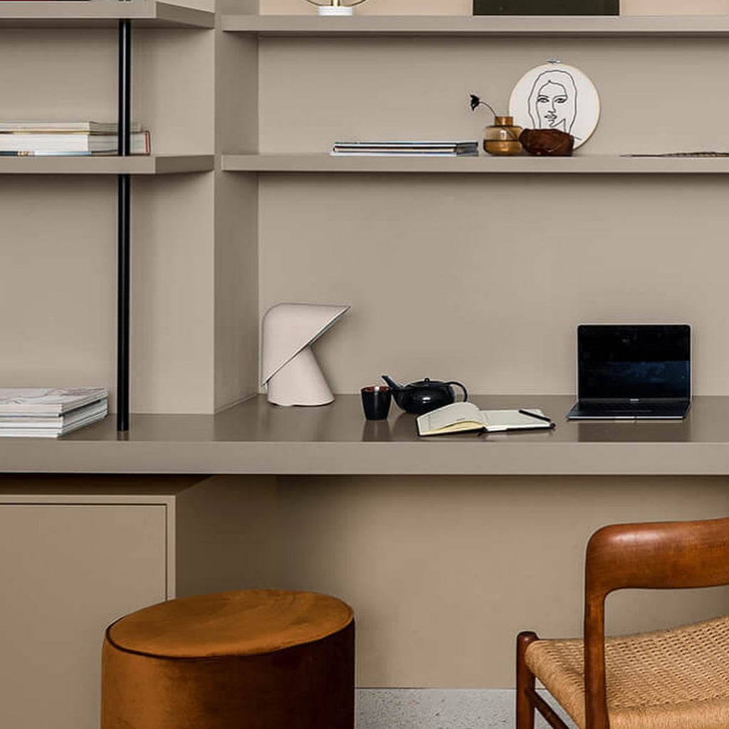 Small white Clérachic table lamp on a taupe shelf desk with a laptop, a teapot and a notebook, beside a leather stool.
