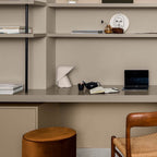Small white Clérachic table lamp on a taupe shelf desk with a laptop, a teapot and a notebook, beside a leather stool.