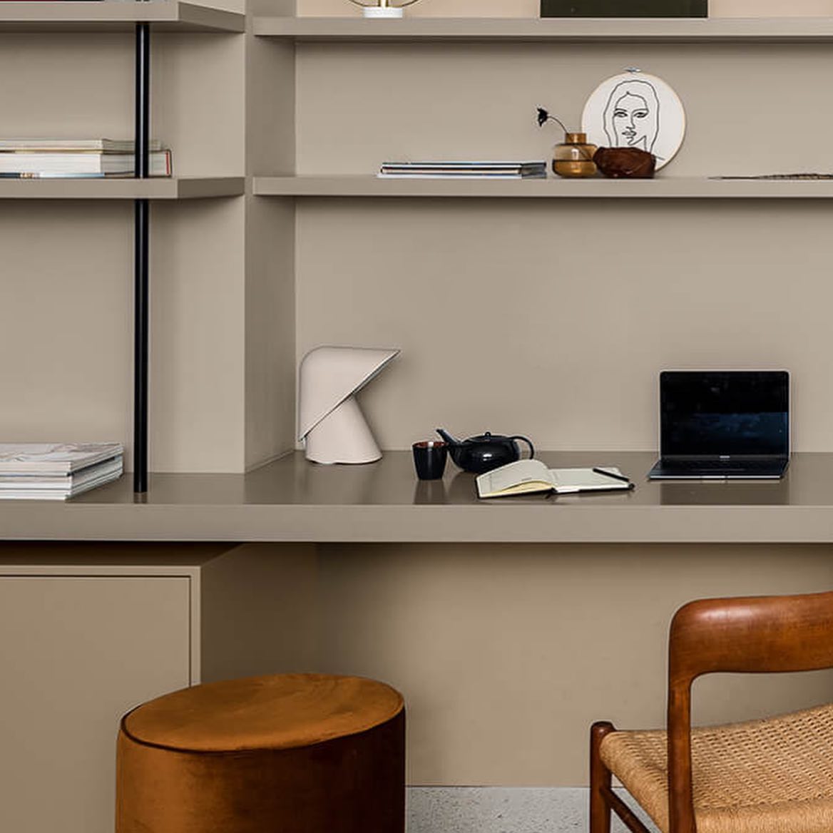 Small white Clérachic table lamp on a taupe shelf desk with a laptop, a teapot and a notebook, beside a leather stool.