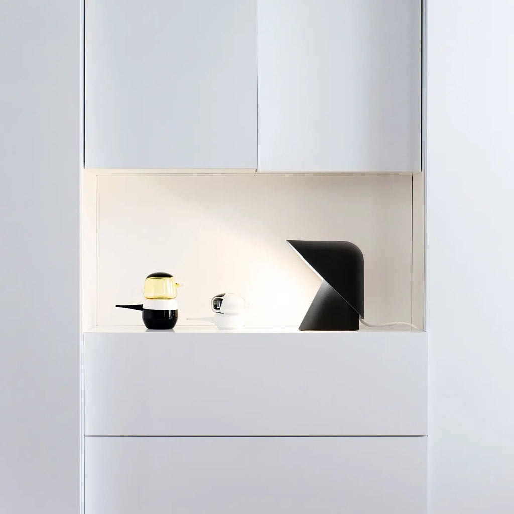 Black Clérachic table lamp in a lit white niche above white drawers, next to a small glass jar and a black-and-white cup.