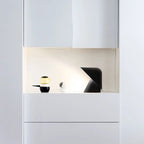Black Clérachic table lamp in a lit white niche above white drawers, next to a small glass jar and a black-and-white cup.