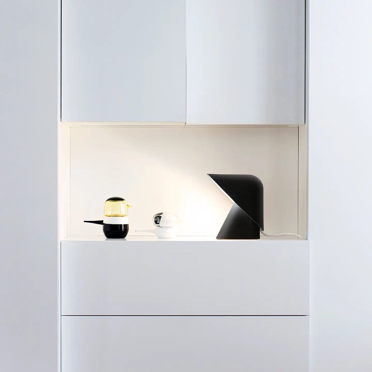 Black Clérachic table lamp in a lit white niche above white drawers, next to a small glass jar and a black-and-white cup.