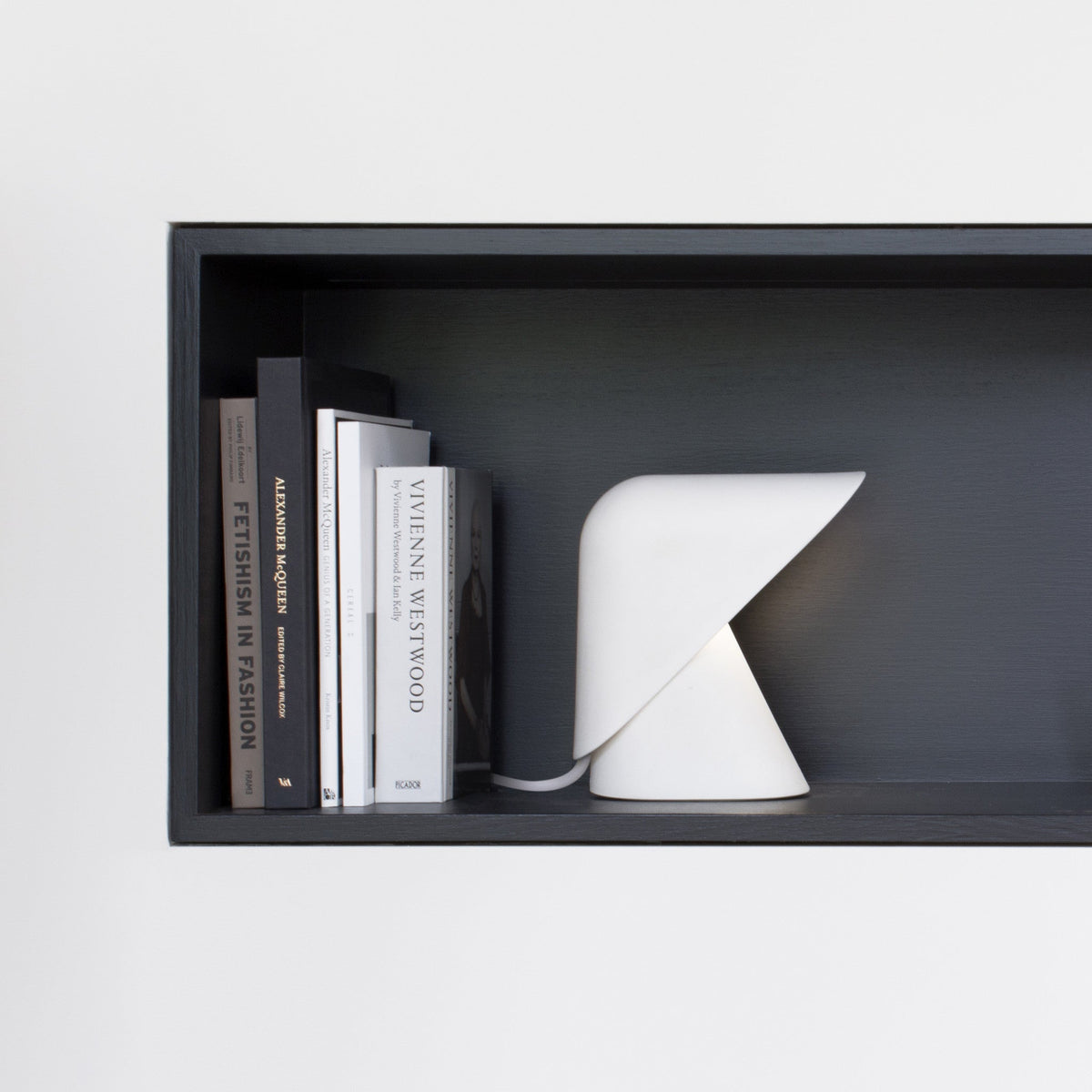 White Clérachic table lamp in a dark shelf recess, standing beside books on fashion and design, against a pale wall.