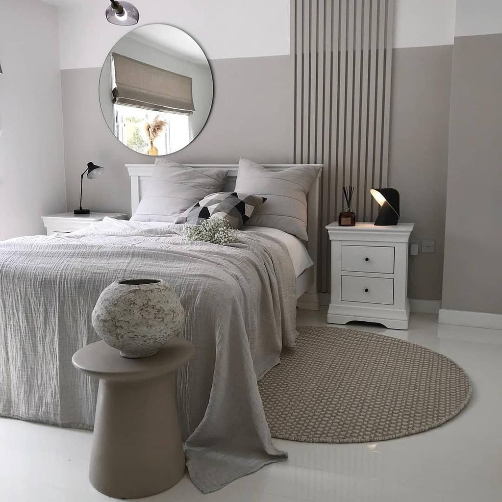 Small black Clérachic table lamp on a white bedside chest in a grey bedroom with a round mirror, a striped panel and a woven rug.