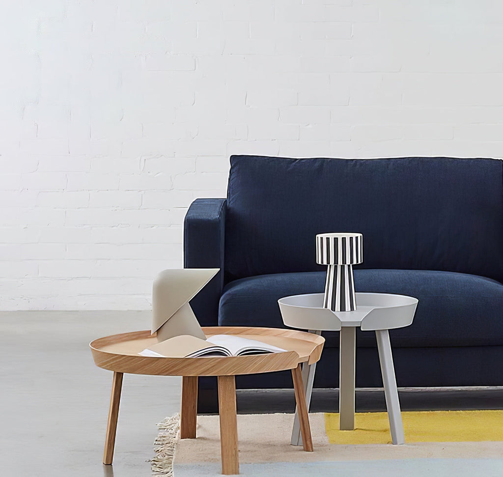 A pale Clérachic table lamp on a round oak coffee table beside an open book, with a navy sofa and a yellow rug behind.