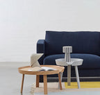 A pale Clérachic table lamp on a round oak coffee table beside an open book, with a navy sofa and a yellow rug behind.