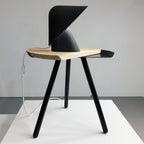 Black Clérachic table lamp on the oak top of a black three-legged side table, with a white cord hanging down the side.