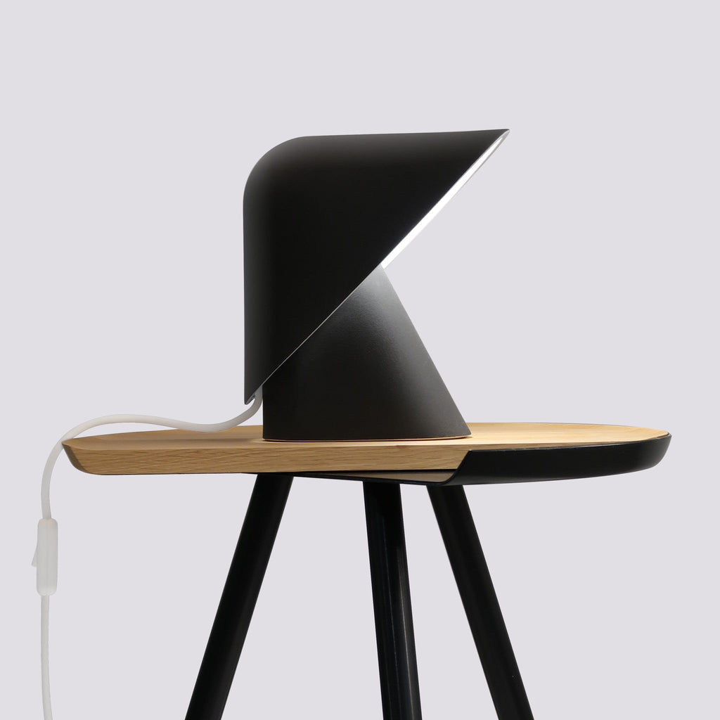 Side view of this Clérachic table lamp in black on a round oak tray table with slim black legs, against a pale grey wall.