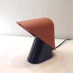 A Clérachic table lamp with an orange shade over a dark navy cone base, on a white surface with a black cord at the back.