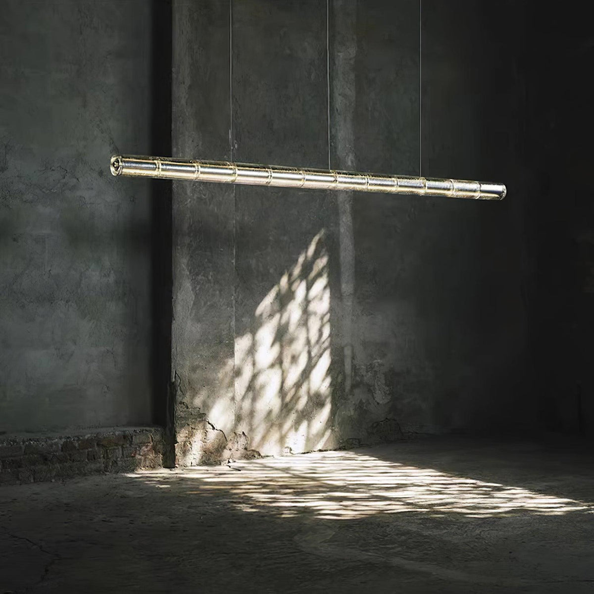 A Clérachic linear chandelier of segmented glass glowing softly in a dark concrete room, with window light patterned on the floor.