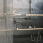 Two glowing segmented glass bars of this Clérachic chandelier hung before a glass wall, above a trestle table with glass vessels.