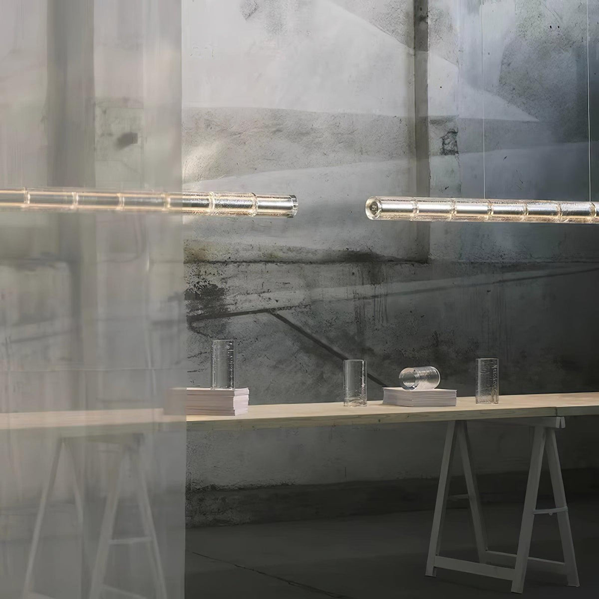 Two glowing segmented glass bars of this Clérachic chandelier hung before a glass wall, above a trestle table with glass vessels.