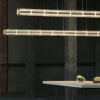 Two warm-lit glass-segment bars of this Clérachic chandelier running across a dark concrete pillar above a table with glass pebbles.