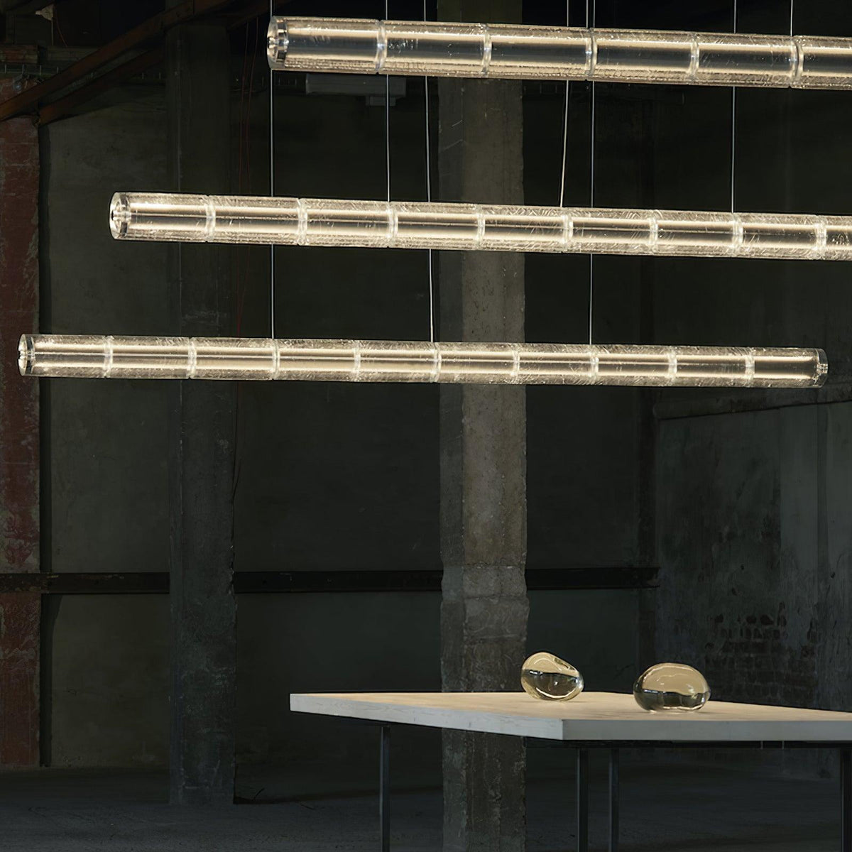 Three Clérachic segmented glass chandeliers glowing warm in a raw industrial hall, with a white table and glass pebbles below.