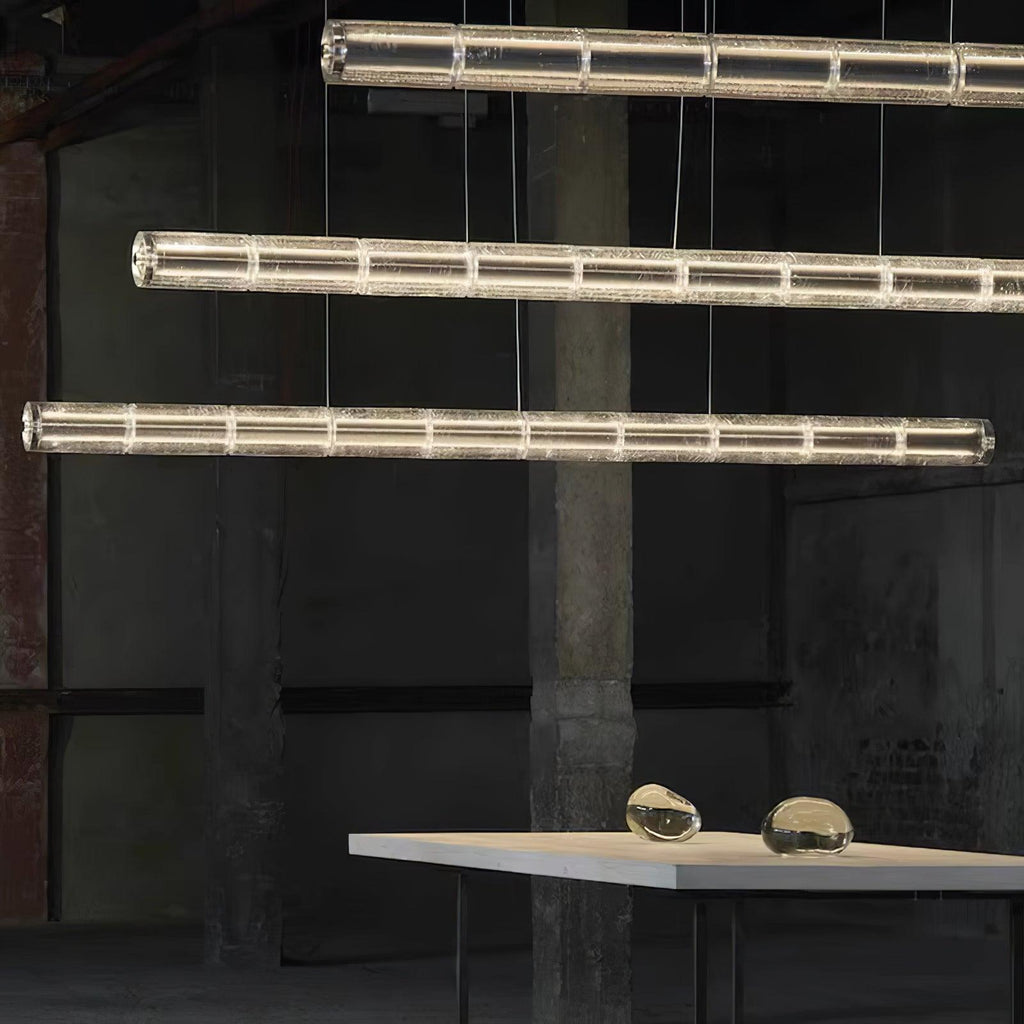 Staggered rows of lit clear glass segments from Clérachic hang beside a concrete pillar, a pale table and two glass pebbles below.