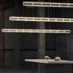 Staggered rows of lit clear glass segments from Clérachic hang beside a concrete pillar, a pale table and two glass pebbles below.