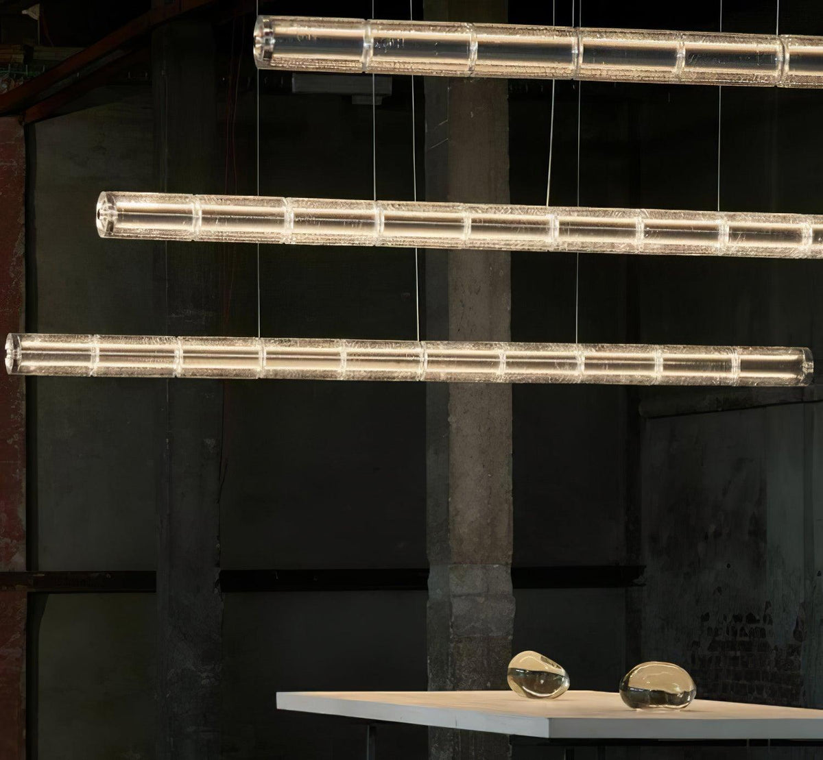Three lit rows of segmented clear glass from Clérachic hang in an industrial space, with a table and glass pebbles at the bottom.