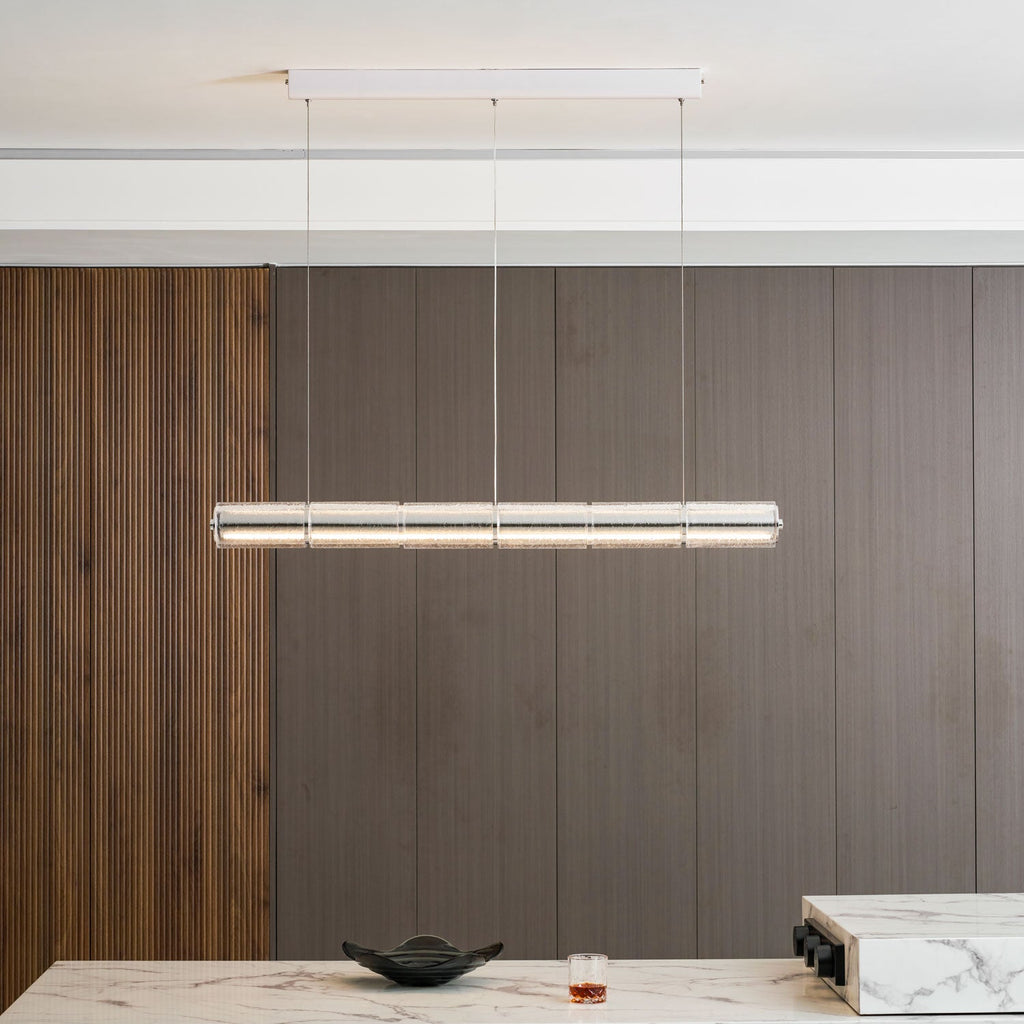A Clérachic linear chandelier of clear glass segments glowing above a marble dining counter, with a dark bowl and a tumbler below.